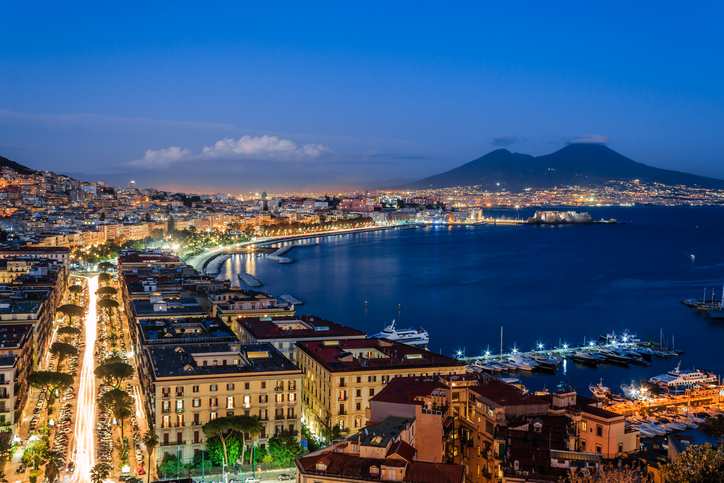 A modern photograph of Mount Vesuvius, viewed across a body of water with a curve of land on the left, similar to the layout of Volaire's painting.