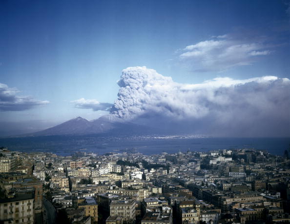 A photograph of the erupting Mount Vesuvius with clouds and smoke in the background and Naples in the foreground. 