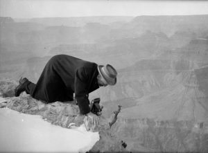 A black and white photograph of Dow on hands and knees at the rocky edge of the Grand Canyon, bending over a camera on the ground in front of him.